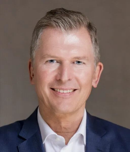 Porträt eines lächelnden Mannes mittleren Alters mit kurzen grauen Haaren, der einen blauen Anzug und ein weisses Hemd trägt vor neutralem Hintergrund.