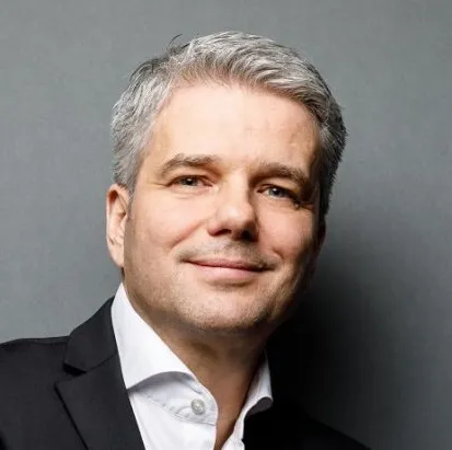 Porträt eines lächelnden Mannes mit grau meliertem Haar in einem schwarzen Anzug und weissem Hemd vor grauem Hintergrund.