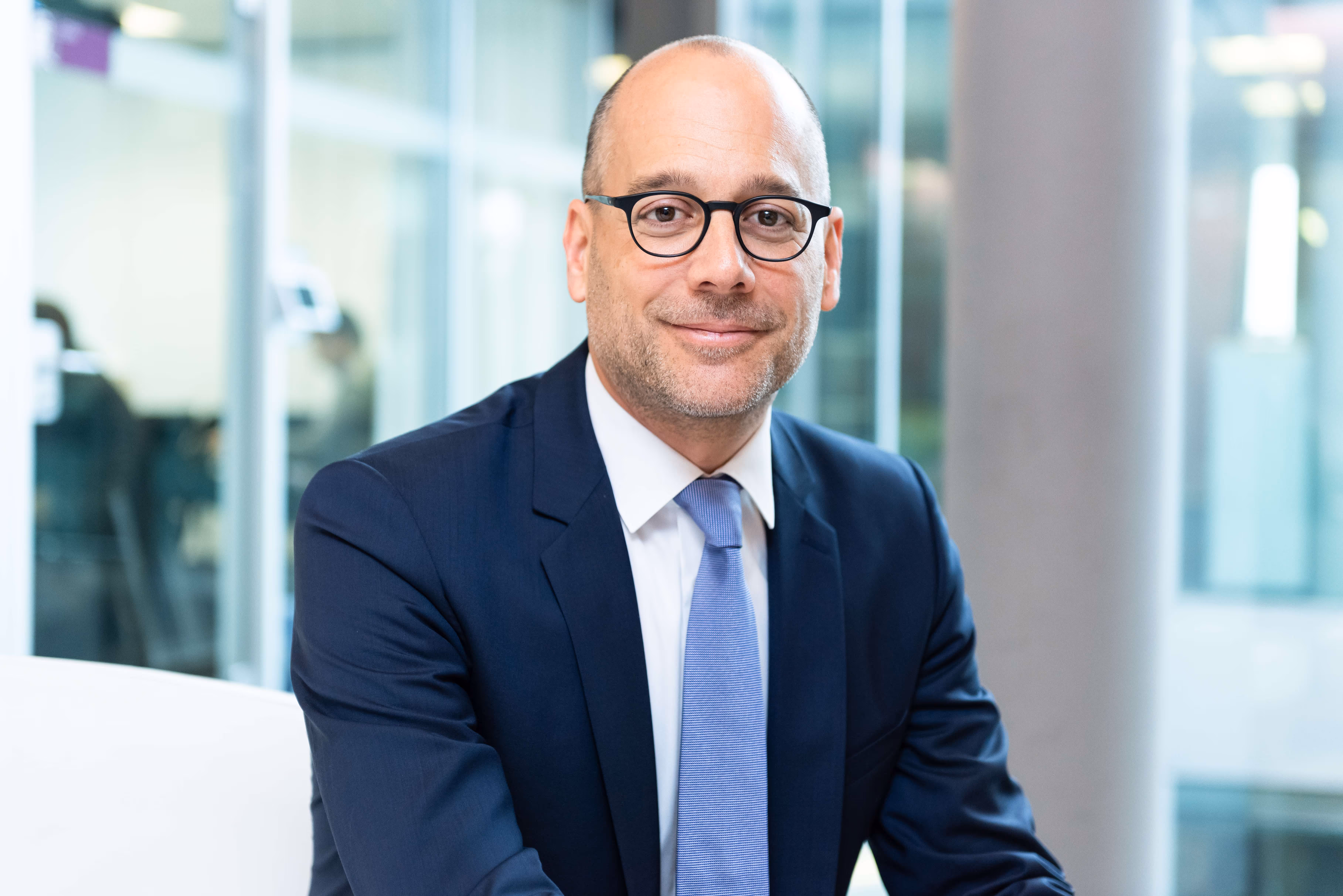 Ein Mann mit Glatze, Brille und blauem Anzug sitzt vor einem modernen Bürohintergrund und lächelt.