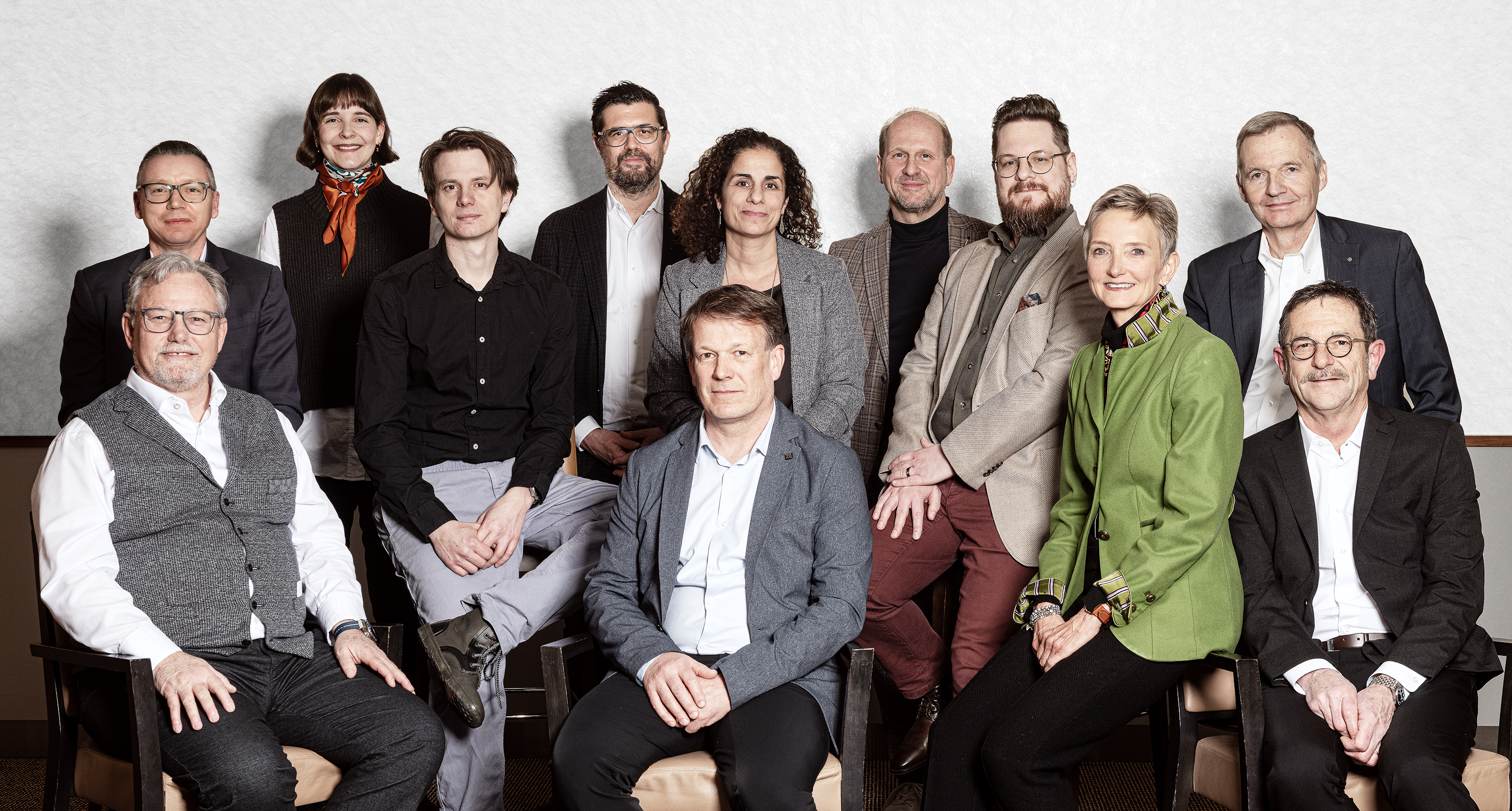 Gruppenfoto von zwölf Personen, elf sitzen und eine steht dahinter, alle tragen formelle Kleidung vor einem hellen Hintergrund.