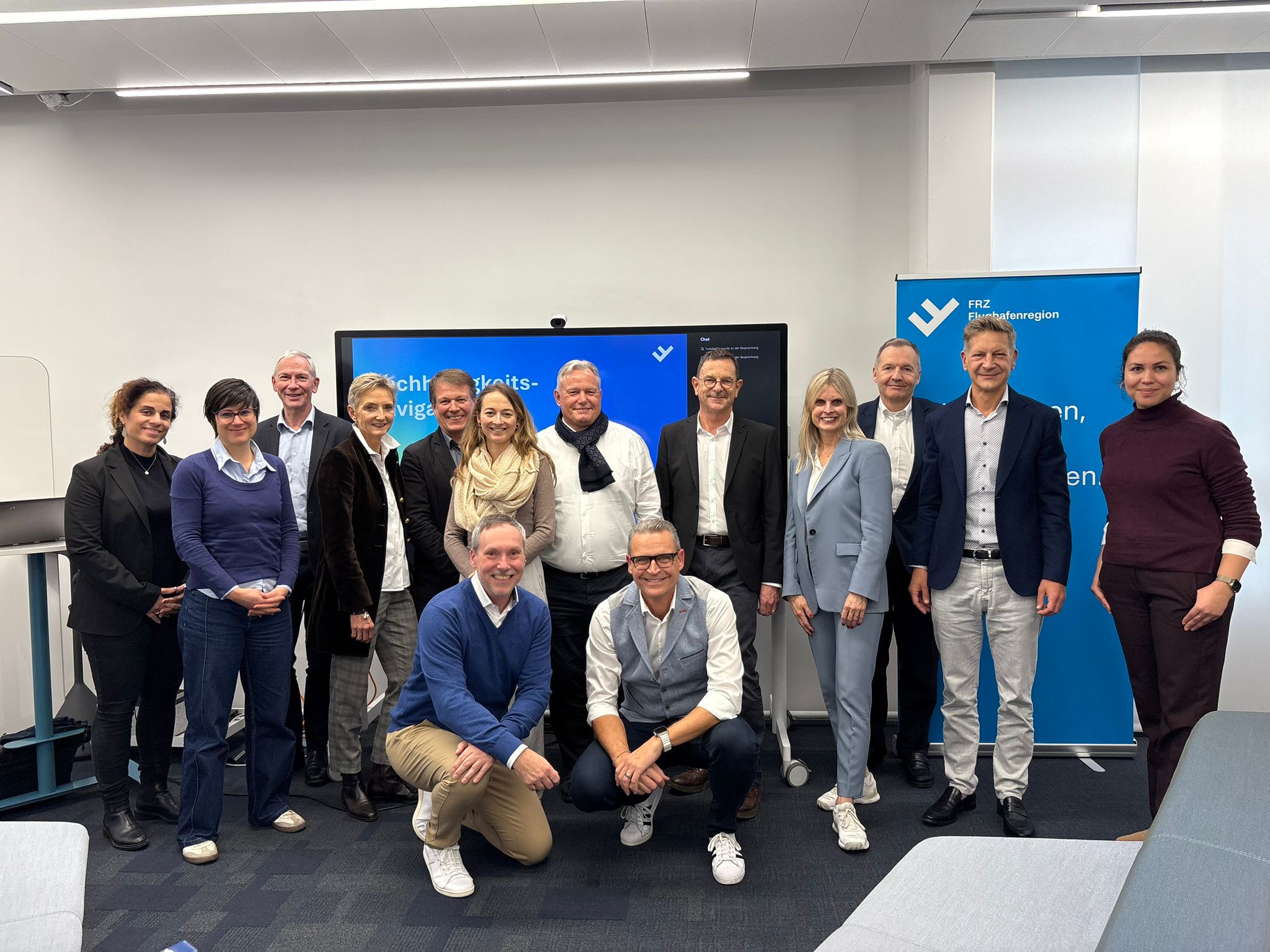 Gruppenfoto von 14 Personen, die in einem modernen Büro vor einem Bildschirm und einem blauen Banner mit dem Logo von FRZ Flughafenregion stehen.