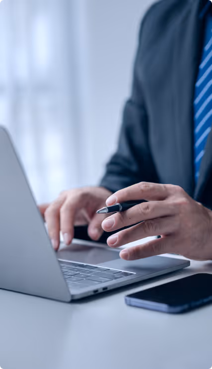Geschäftsmann tippt auf Laptop, hält Kugelschreiber, Smartphone liegt auf Tisch.