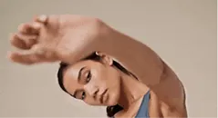 Woman with long hair in a blue top raising her arm covering part of her face.