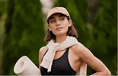 Woman in tank top and cap holding a yoga mat outdoors with greenery in background.