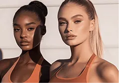 Two women with makeup and ponytails wearing orange tops posing against a light background.