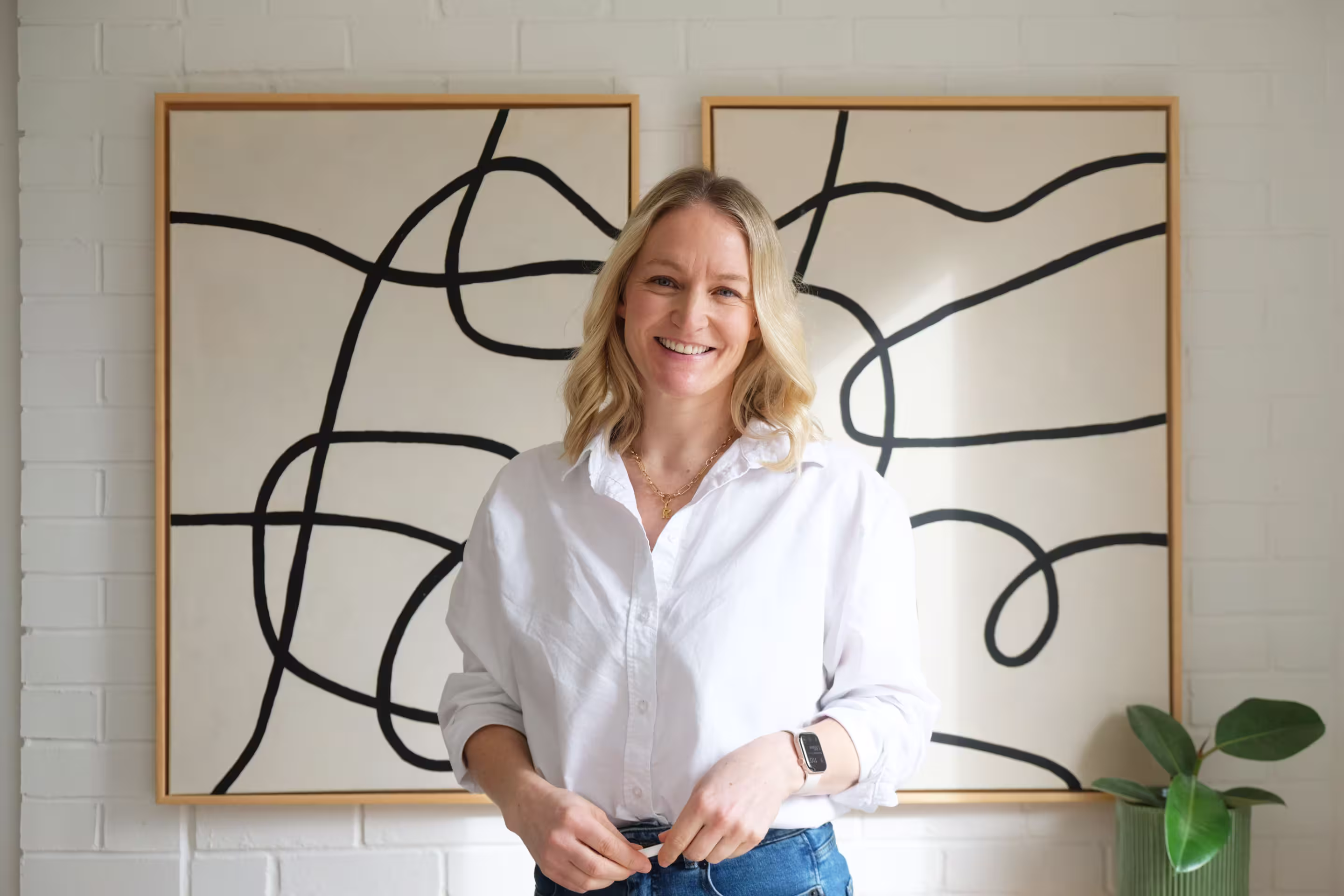 Smiling woman with blonde hair wearing a white shirt and blue jeans standing in front of two abstract paintings with black lines on a white background.