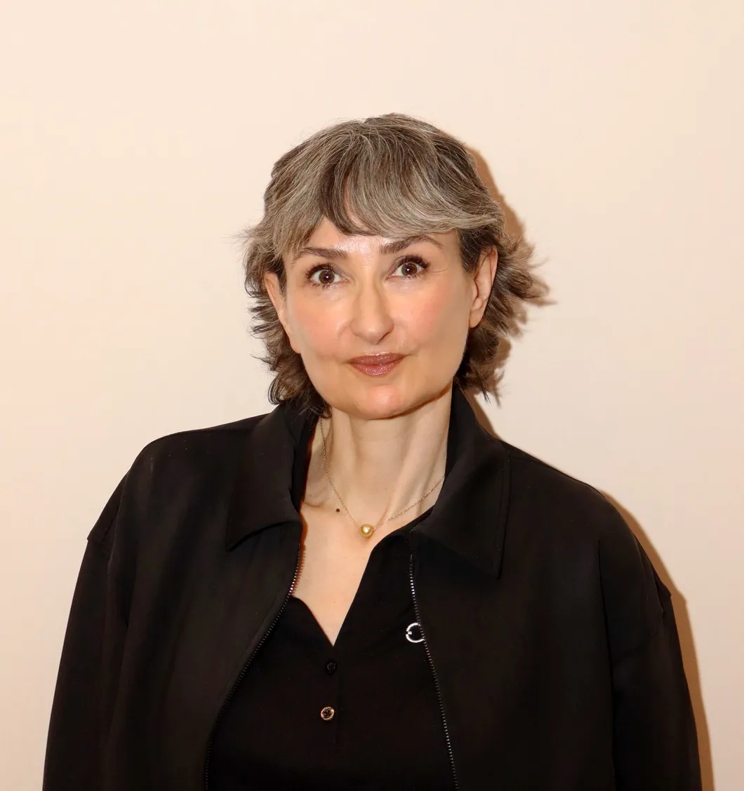 Woman with short gray hair wearing a black jacket and a pearl necklace against a plain background.