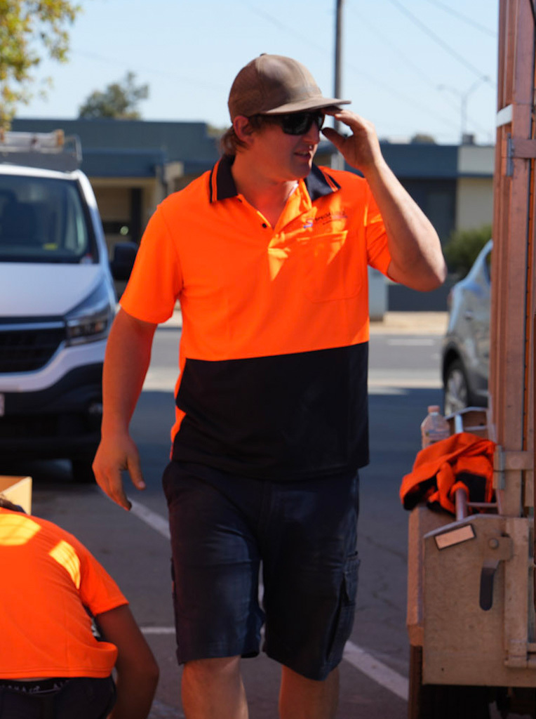 Man in orange and black work shirt, wearing cap and sunglasses, walking outdoors near a truck.