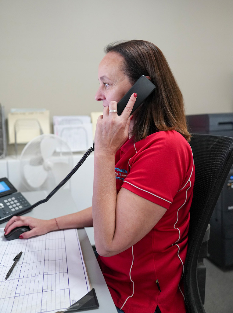 Woman in red shirt sitting at desk using telephone and computer mouse.