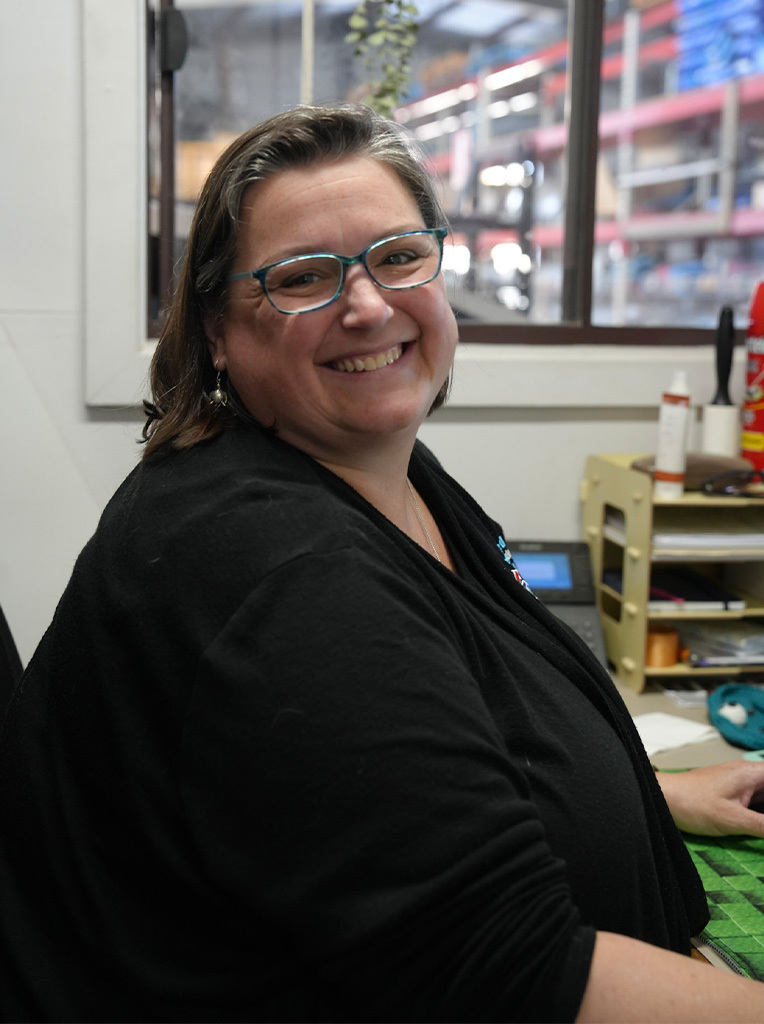 Smiling woman wearing glasses sitting at desk with shelves and window behind her.