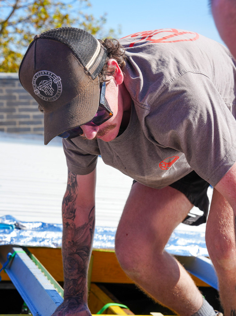 Man with tattoos wearing a cap and sunglasses working outdoors on a construction or repair project.