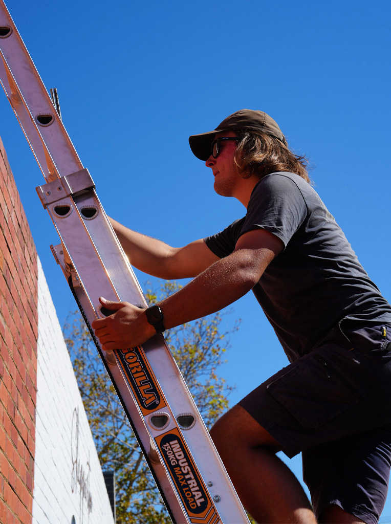 Man wearing cap and sunglasses climbing an industrial aluminum ladder against a brick wall.