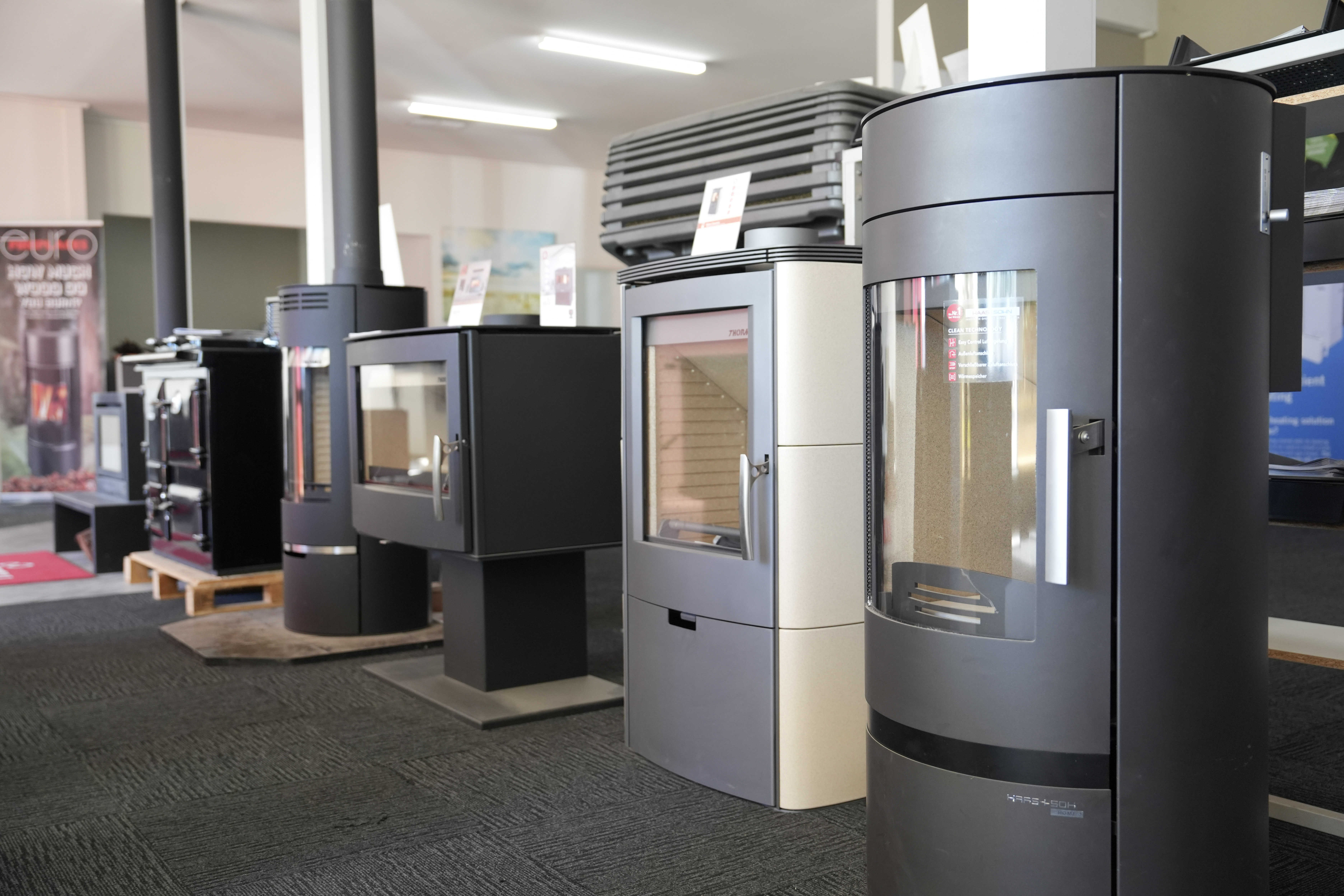 Several modern wood-burning stoves on display in a showroom with carpeted floors and bright lighting.