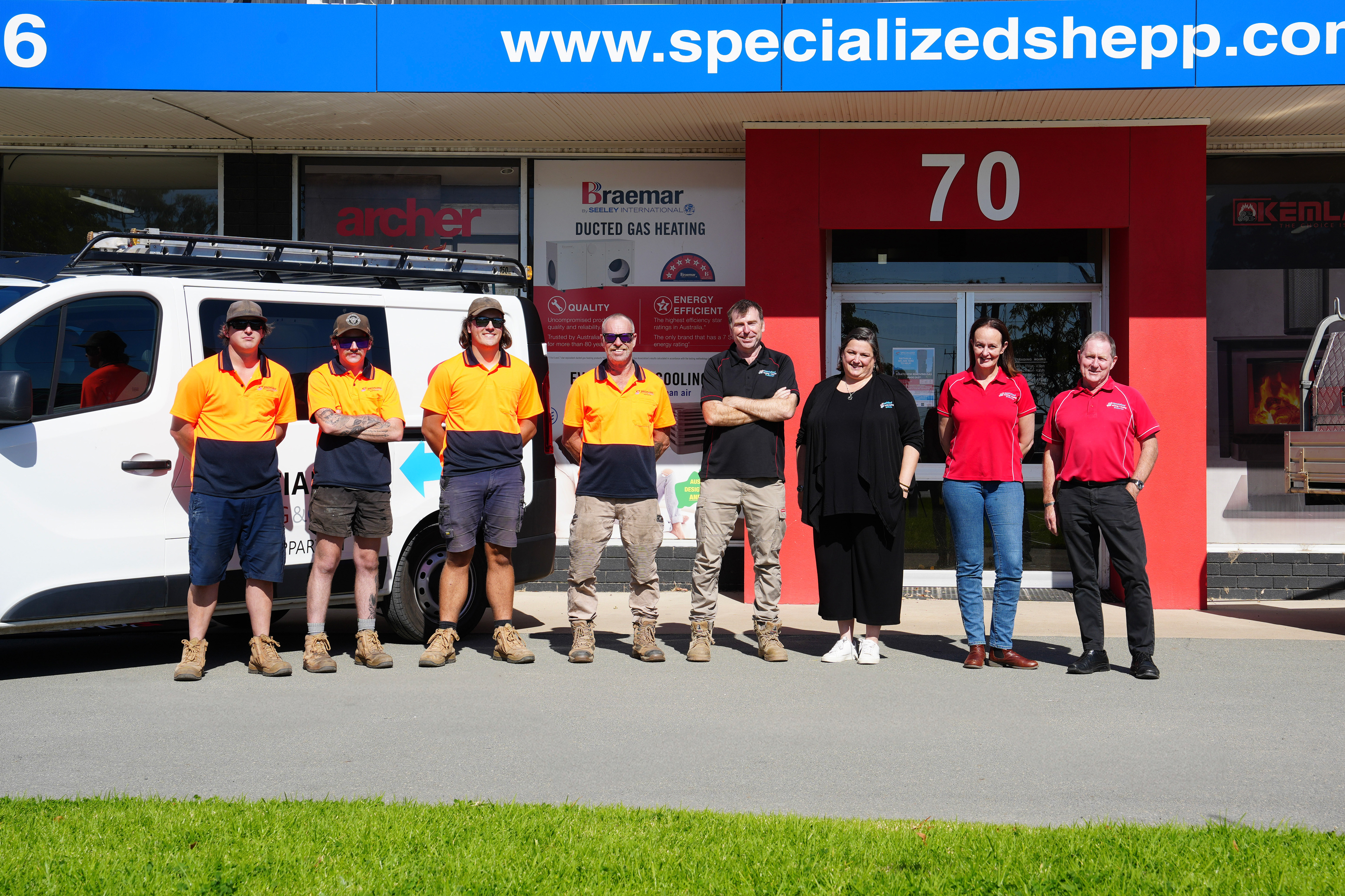 Eight workers standing in a row outside a building with red walls and the number 70 above the door.