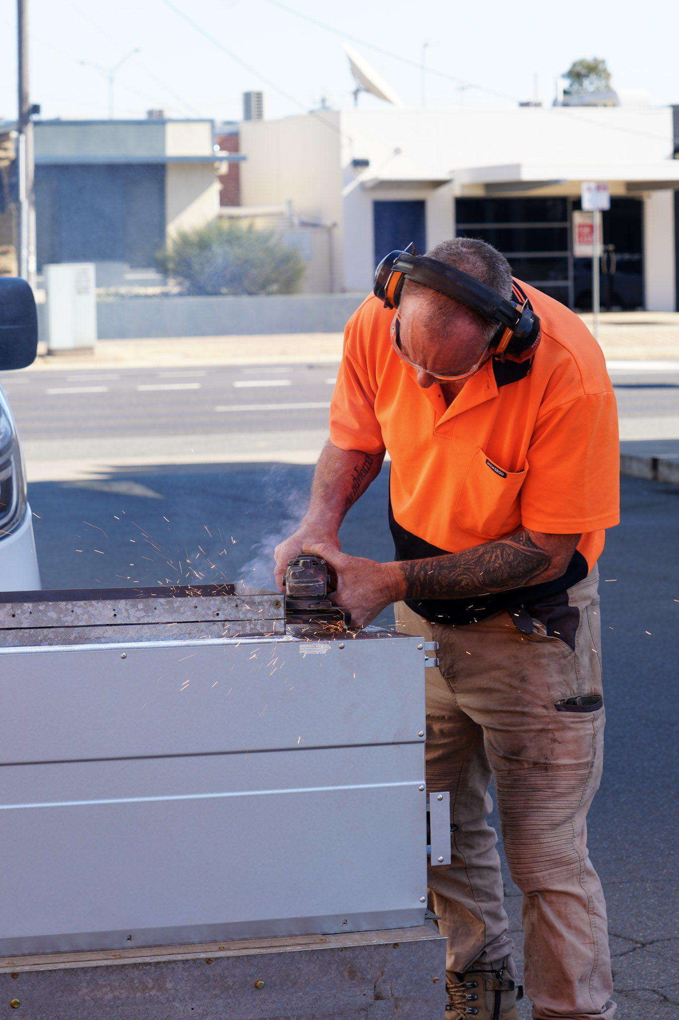 Man in orange shirt using a power tool on metal outdoors with sparks and smoke visible.