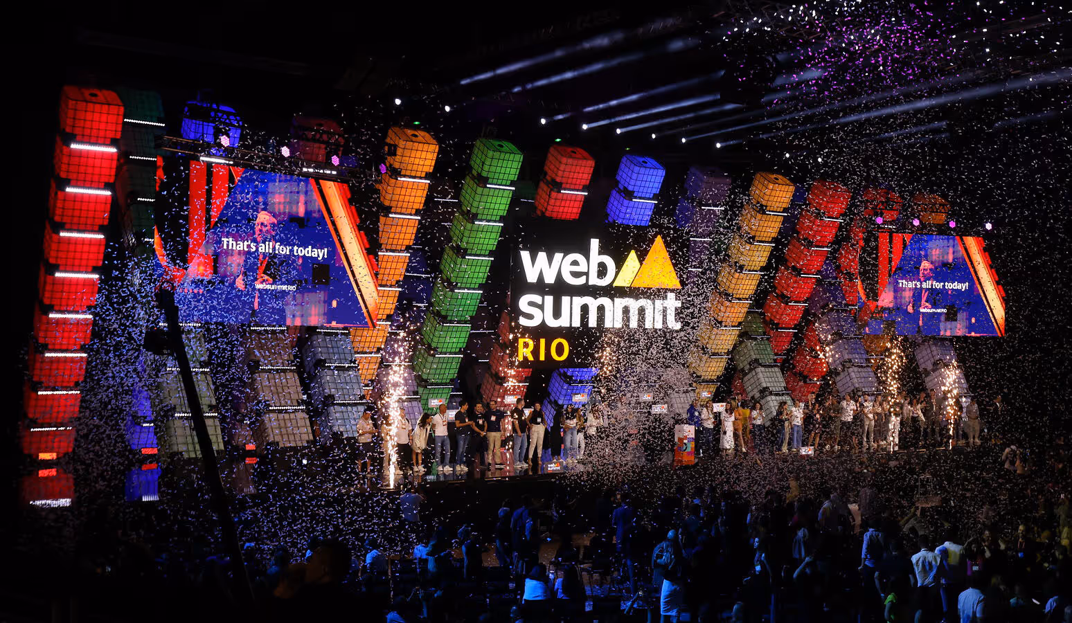 A large stage viewed over the silhouetted heads of a packed audience. A line of people stands on the stage, facing towards the audience. Confetti fills the air. A large Web Summit Rio logo hangs in the center of the stage. Two screens on either side of the logo read 'That's all for today!'