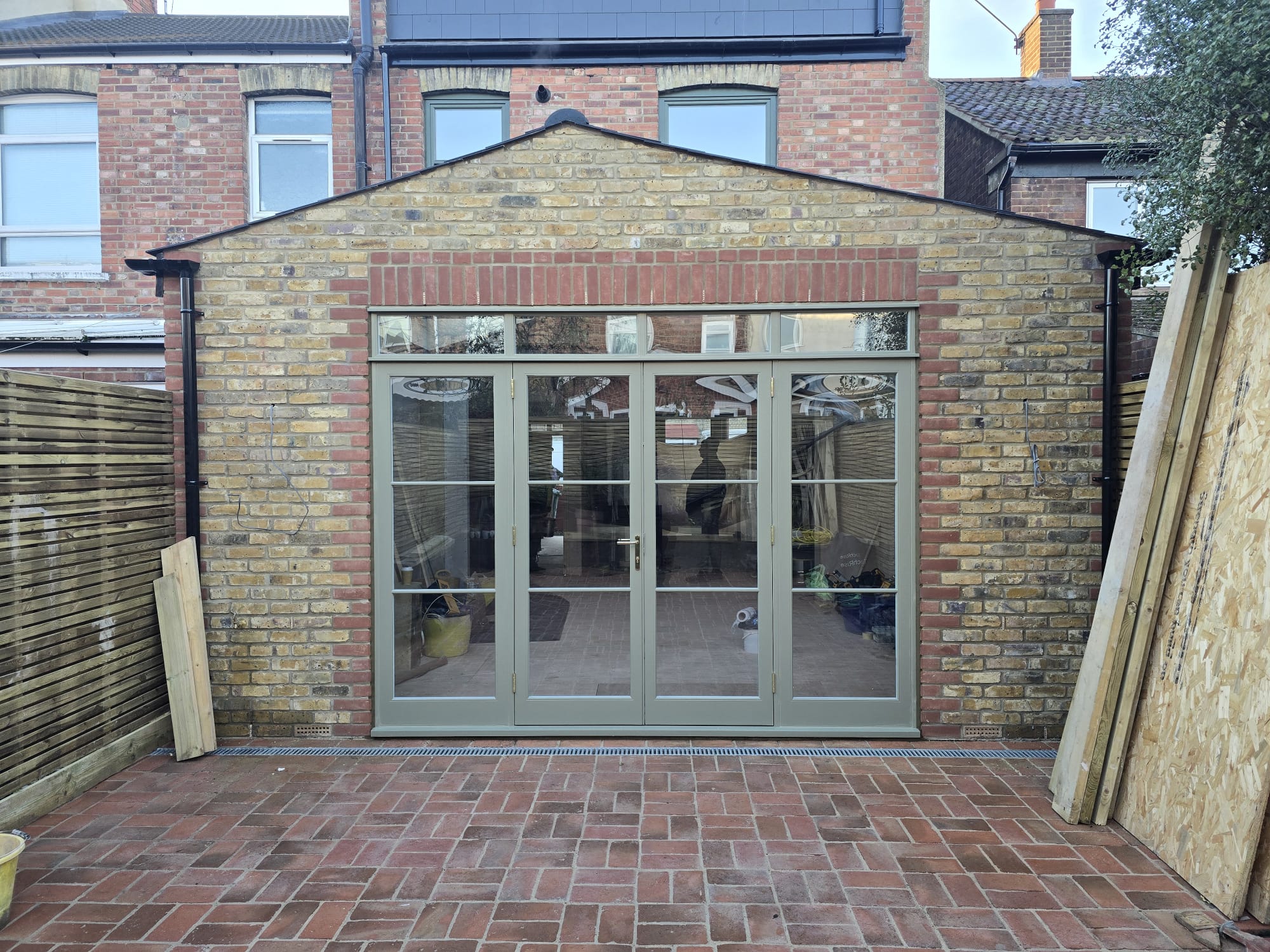 Sage green timber bi-fold doors with glazed transom on brick garden extension by AMB Joinery