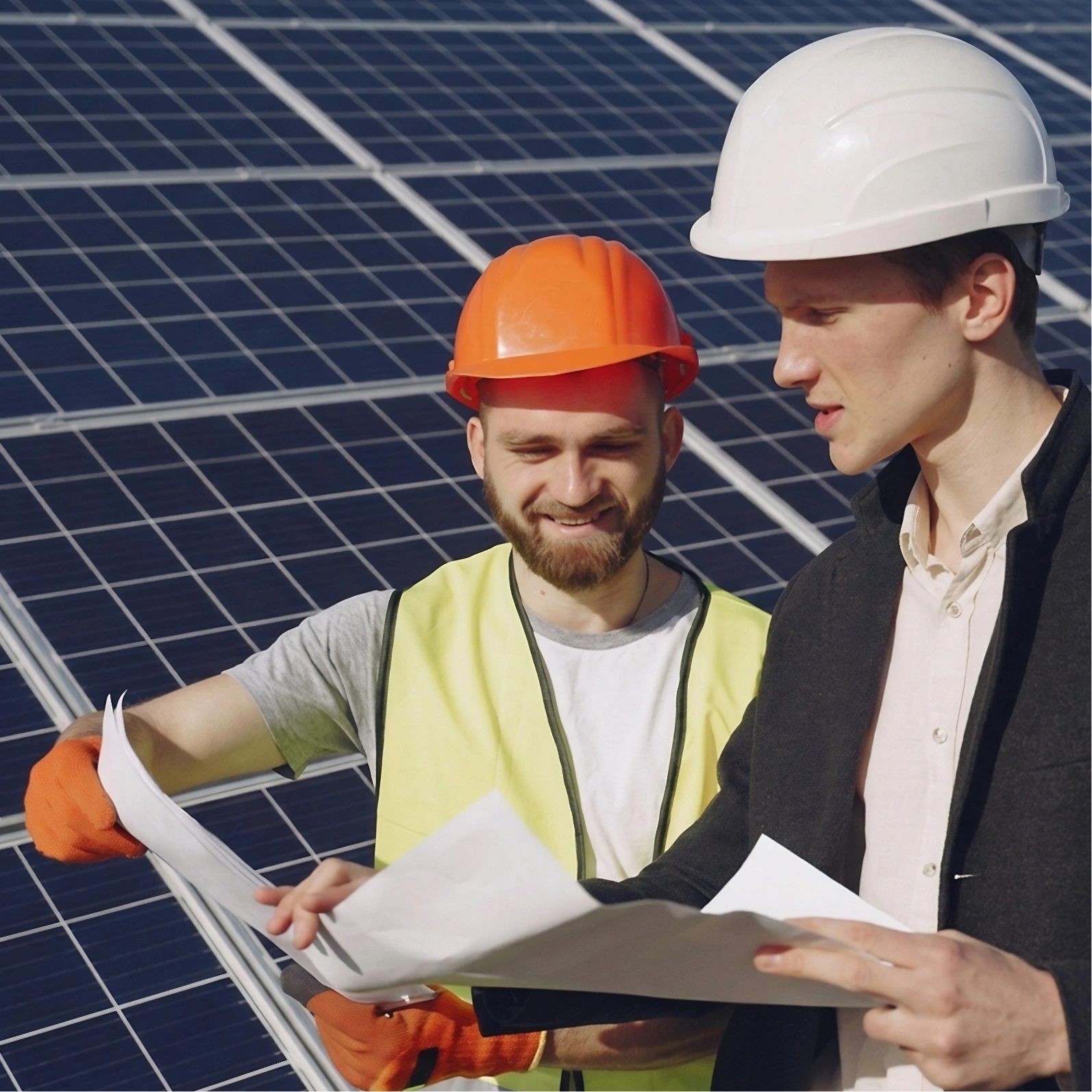 Two male workers wearing safety helmets review plans in front of solar panels.