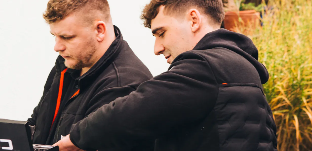 Zwei Männer in schwarzen Jacken arbeiten gemeinsam im Freien an einer Aufgabe.