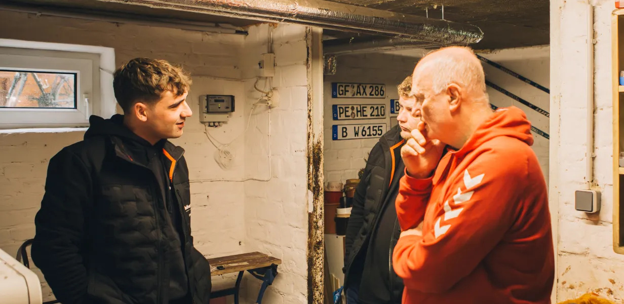 Zwei Männer in schwarzen Jacken sprechen mit einem Mann in orangem Hoodie in einer Werkstatt mit weißen Backsteinwänden und Kfz-Kennzeichen an der Wand.