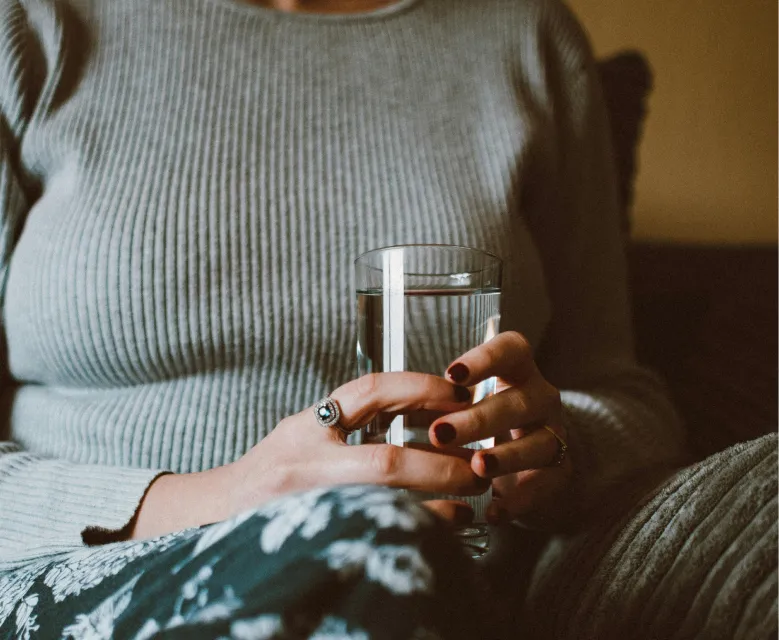 Person in grauem Rippenpullover hält mit beiden Händen ein klares Glas Wasser, Hose mit Blumenmuster sichtbar.