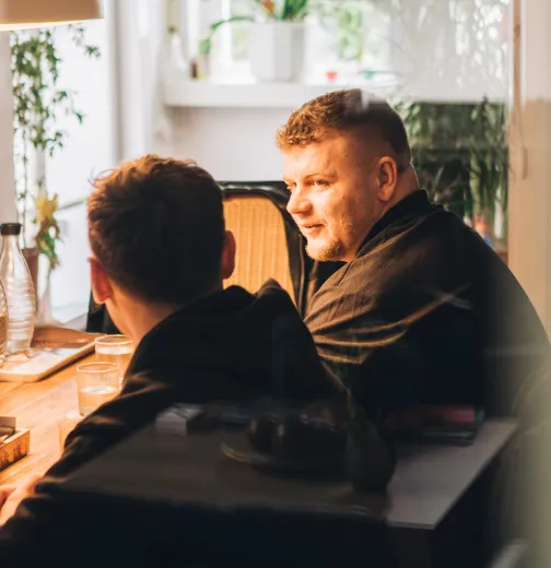 Zwei Männer sitzen an einem Holztisch am Fenster und unterhalten sich in einem hellen Raum mit Pflanzen.