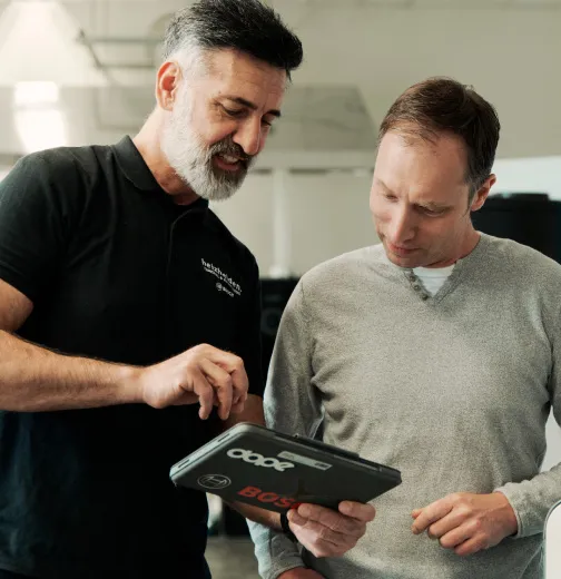 Techniker in schwarzem Shirt zeigt einem Mann in grauem Pullover Informationen auf einem Tablet.