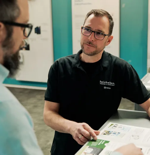 Mann in schwarzem Shirt und Brille erklärt einem bärtigen Mann mit Brille Dokumente in einem Büro.
