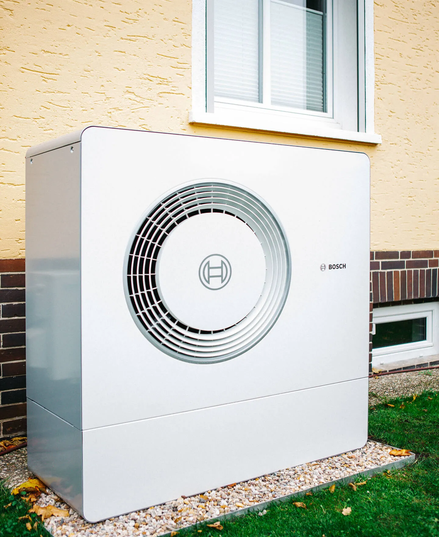Bosch-Außenwärmepumpe auf Kies neben einer gelben Hauswand mit Fenster installiert.