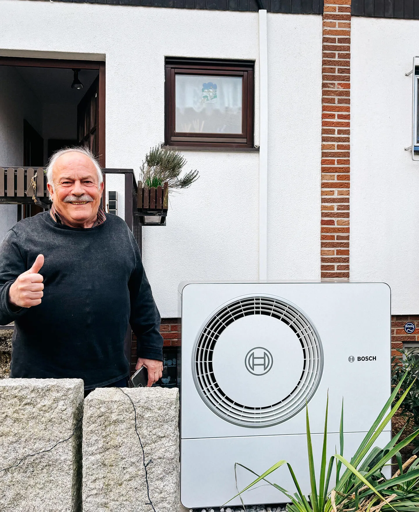 Lächelnder älterer Mann zeigt Daumen hoch neben einer Bosch-Außenwärmepumpe vor einem Haus.
