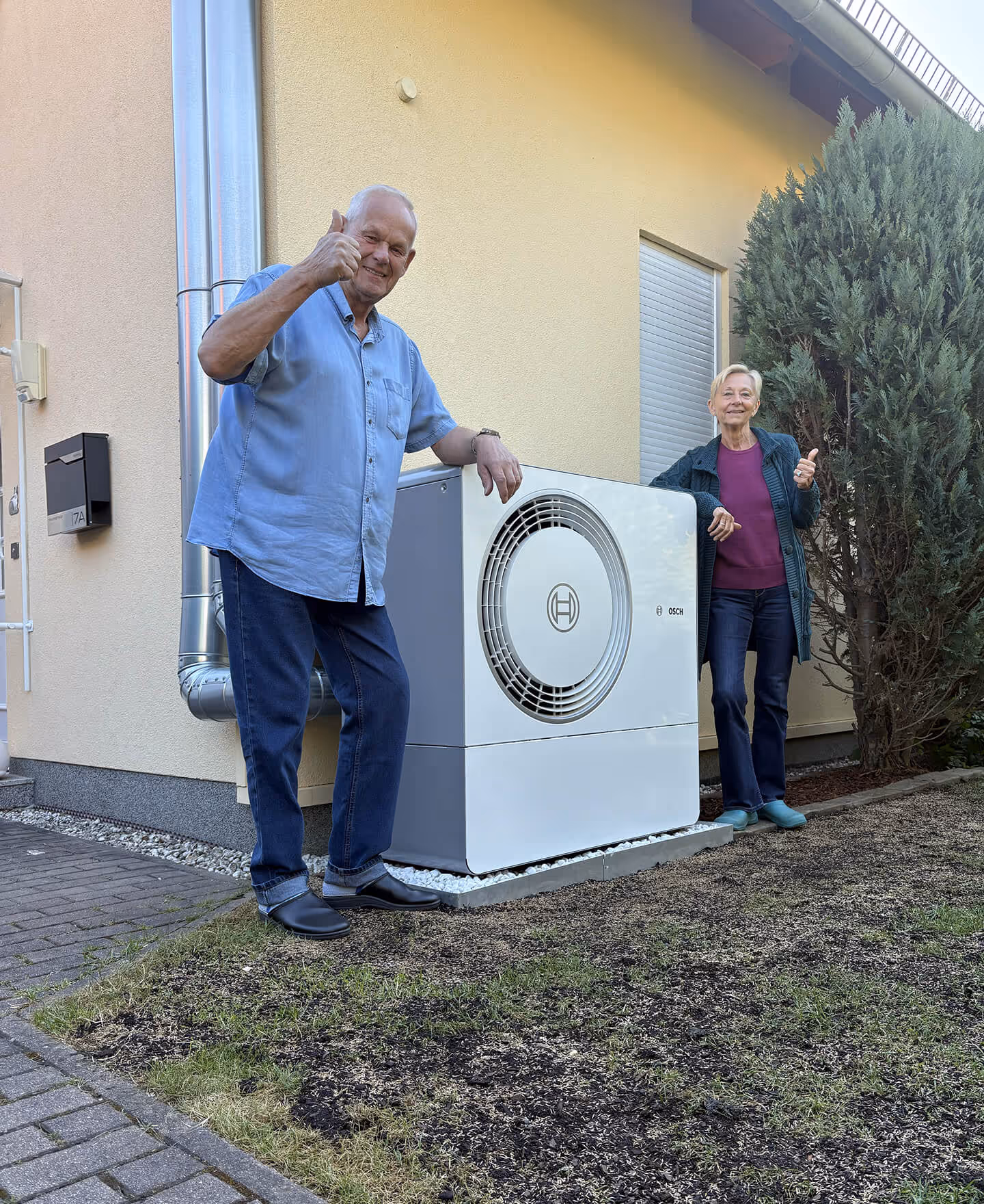 Älterer Mann und ältere Frau stehen vor einem Haus neben einer großen Bosch-Wärmepumpe und zeigen beide Daumen hoch.