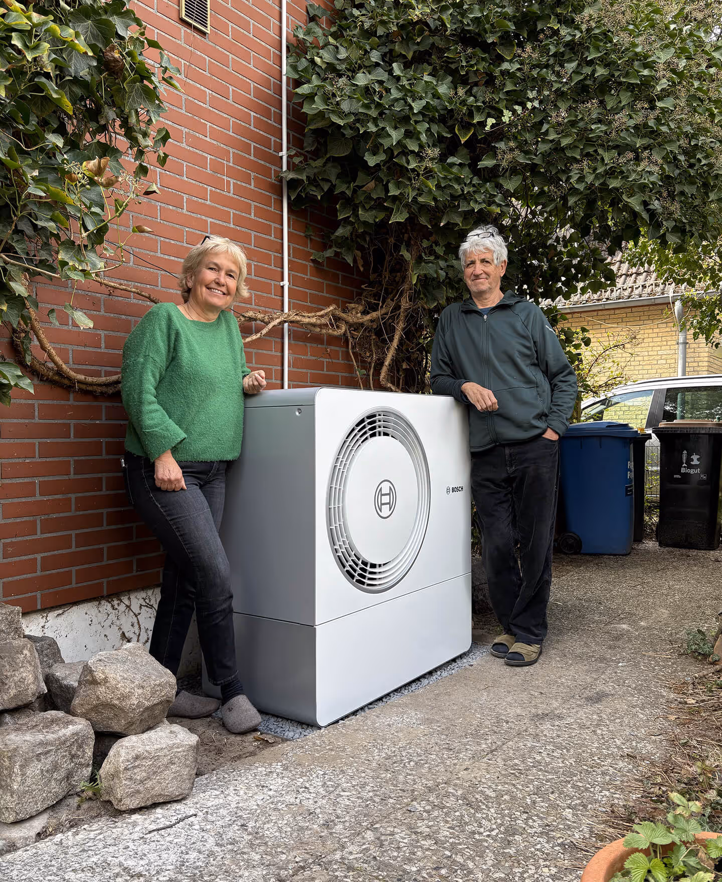 Zwei Personen stehen neben einer großen weißen Bosch-Wärmepumpe im Freien neben einer Backsteinwand mit Blattpflanzen.