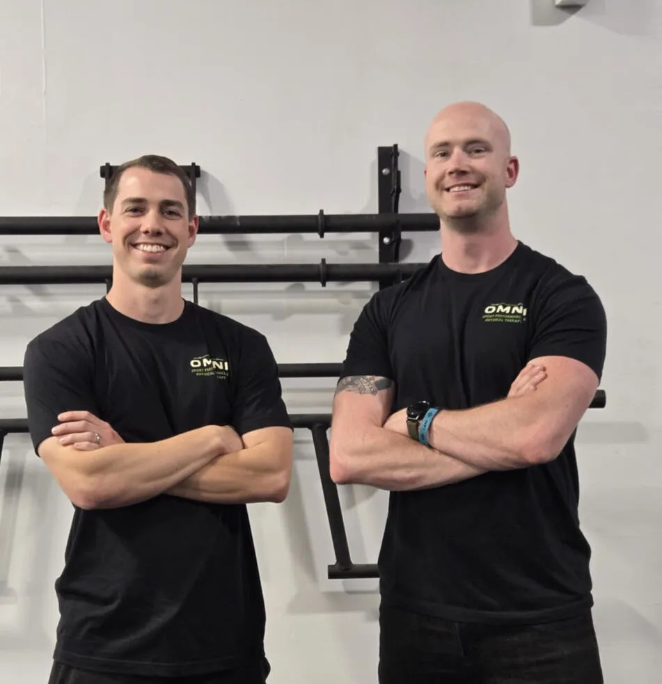Two men smiling with arms crossed, wearing black t-shirts with Omni Physical Therapy logo standing indoors.
