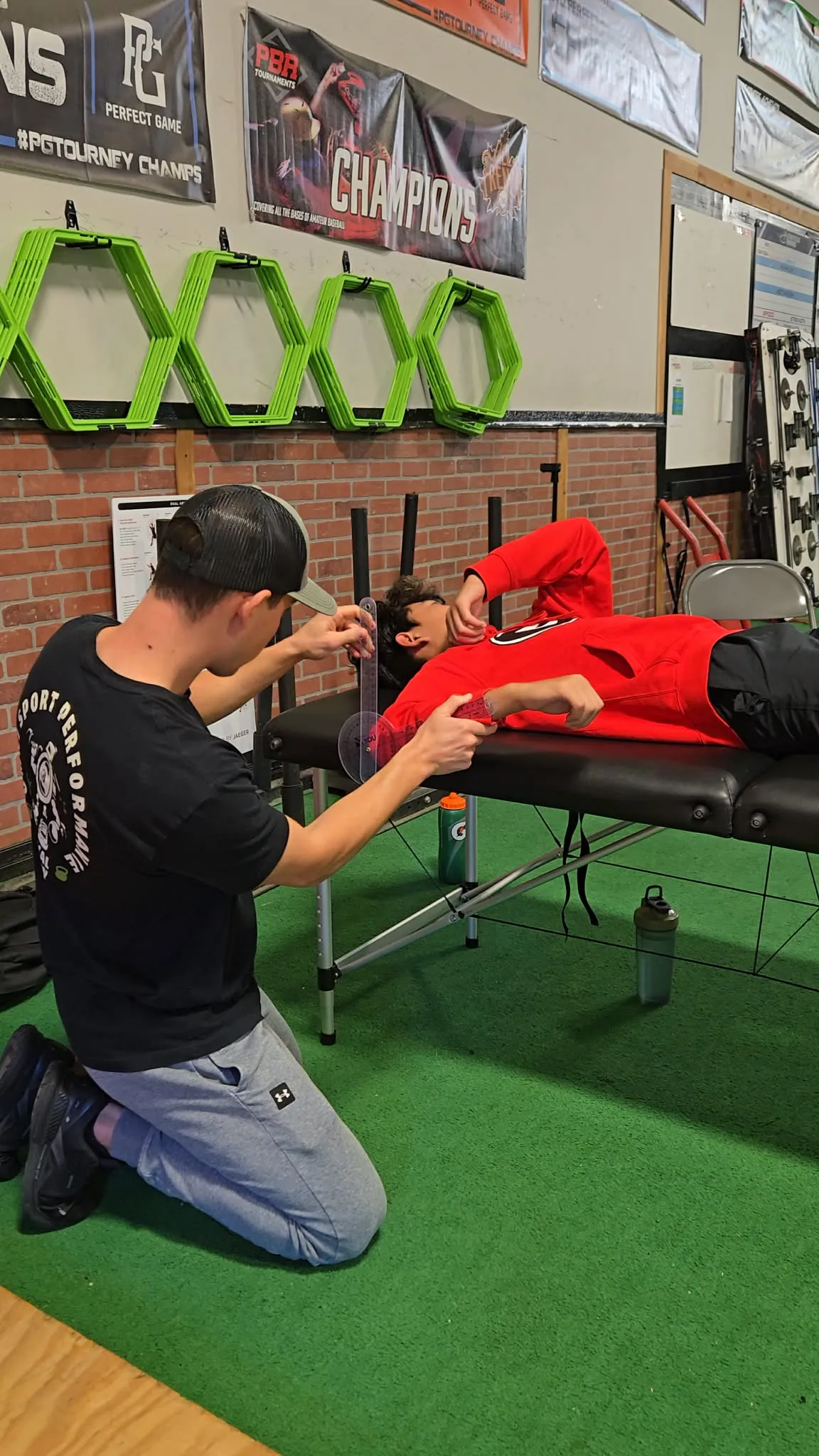 Trainer kneels measuring arm angle of a person lying on a black massage table in a gym.