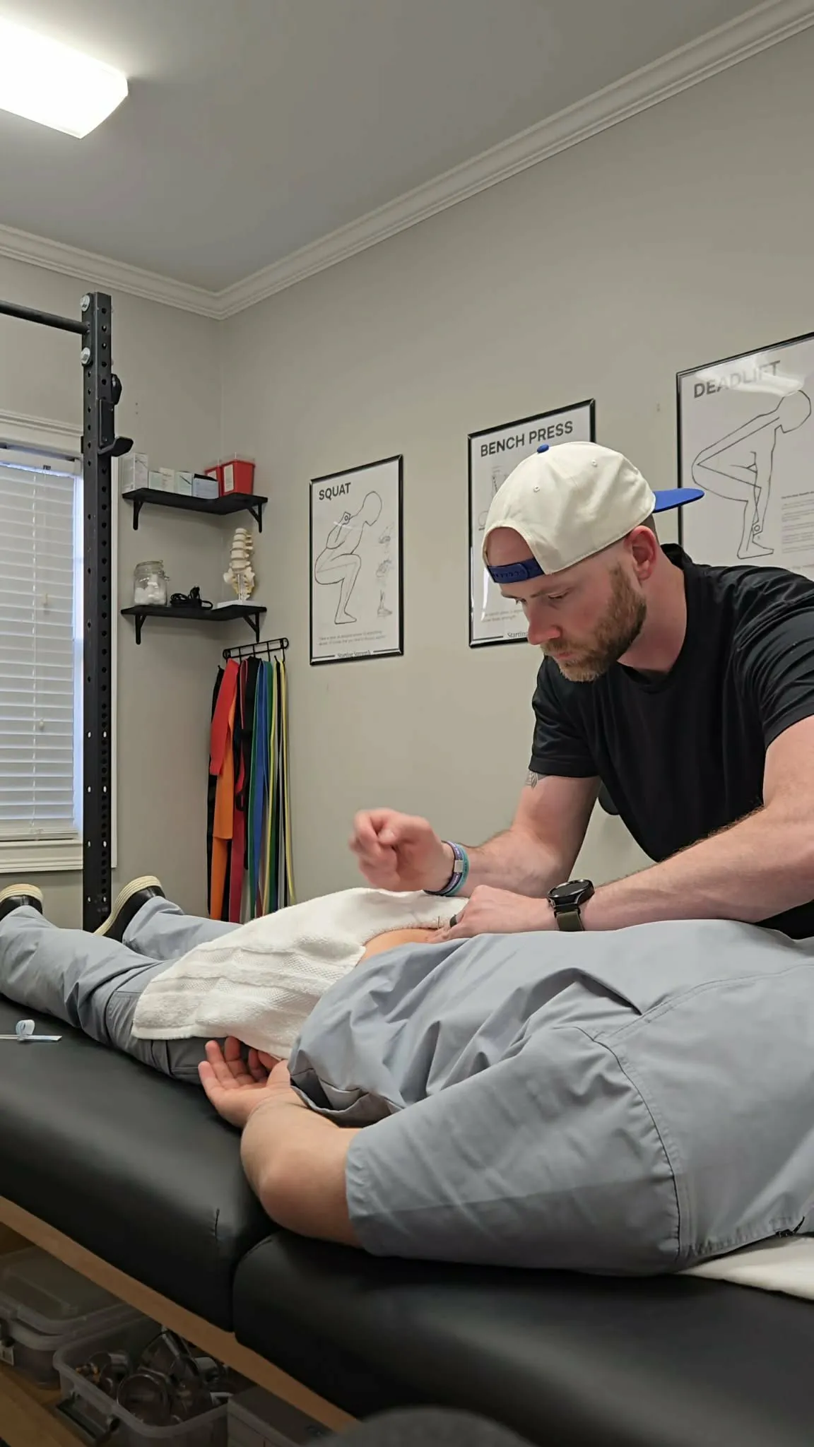 Man wearing a backward cap performs acupuncture on a person lying face down on a treatment table.