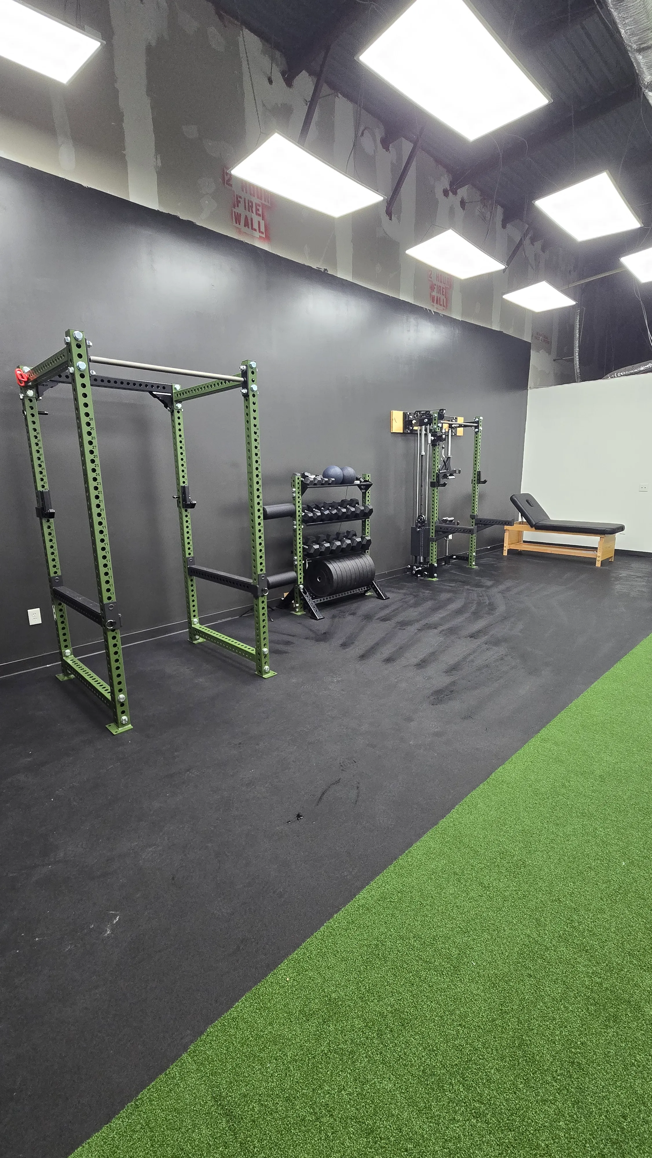 Minimal gym corner with green power rack, dumbbells, weight plates, cable machine, and incline bench on artificial turf.
