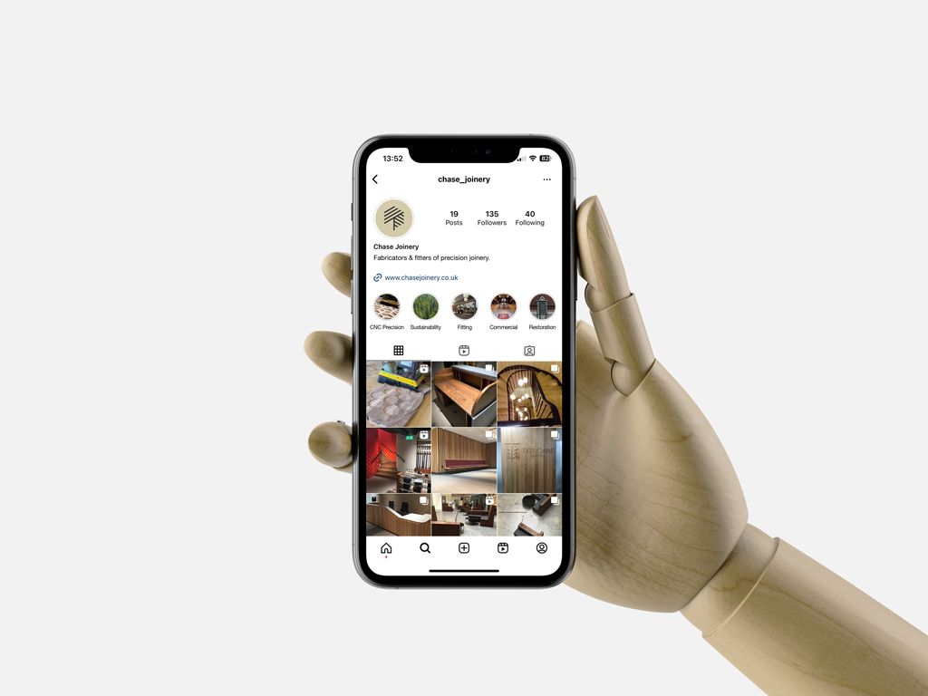 Wooden mannequin hand holding a phone displaying the Instagram page of Chase Joinery with posts showcasing woodwork projects.