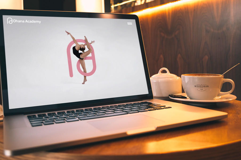 Laptop on wooden table displaying Ohana Academy website with a dancer illustration, next to a cup of coffee and a sugar bowl.