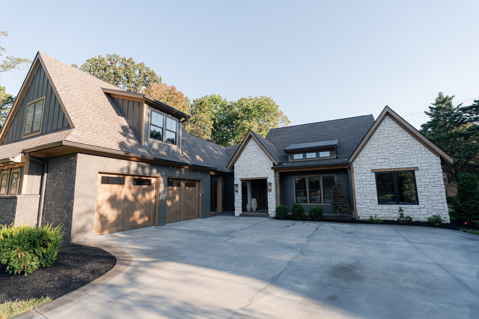 Modern house exterior with mixed dark gray brick and white stone walls, two-car wooden garage doors, large windows, and a spacious concrete driveway surrounded by landscaped bushes and trees.