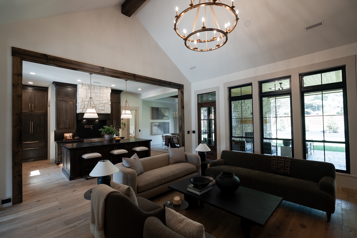 Modern open-concept living room and kitchen with high vaulted ceilings featuring a large circular chandelier, a beige sofa with pillows, a black sofa, armchairs, a black coffee table with decorative items, and large windows with black frames overlooking a patio with stone walls. The kitchen has dark wood cabinetry, a stone range hood, a black countertop island with three cushioned stools, and pendant lights hanging above the island.