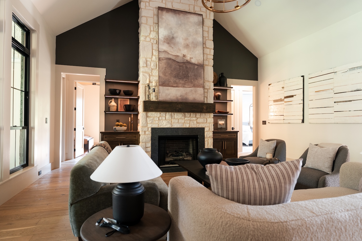 Modern living room with a stone fireplace centered between two dark wood cabinets with shelves, decorated with pottery and candles, light beige and gray armchairs and sofas facing a black coffee table, wooden floors, and large windows letting in natural light.