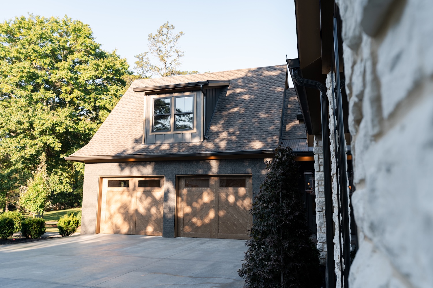 Modern house exterior with a dual wooden garage door, gray brick walls, and a dormer window on the roof, surrounded by greenery.