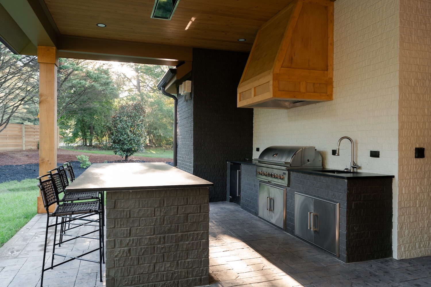 Modern covered outdoor kitchen with black brick counter, stainless steel grill, sink with faucet, wooden range hood, and three high metal chairs along the beige-topped island, surrounded by greenery.