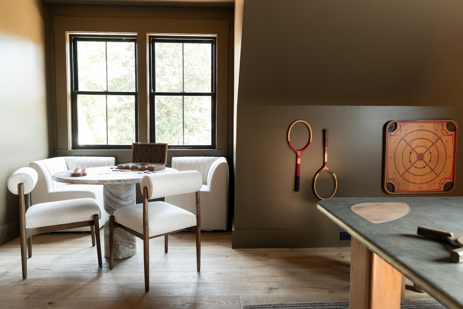 Cozy room with light wooden floor and olive walls featuring a round marble table surrounded by cushioned chairs and a curved sofa near two windows, vintage wooden tennis rackets hanging on the wall, and a wooden board game mounted beside them.