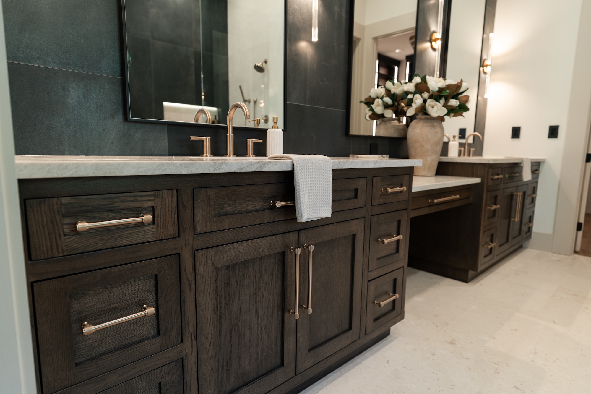 Modern bathroom with dark wood cabinetry featuring metal handles, a white countertop with gold faucets, a hanging white towel, and a large beige vase filled with white flowers on the right side against a black tiled wall with a large mirror.