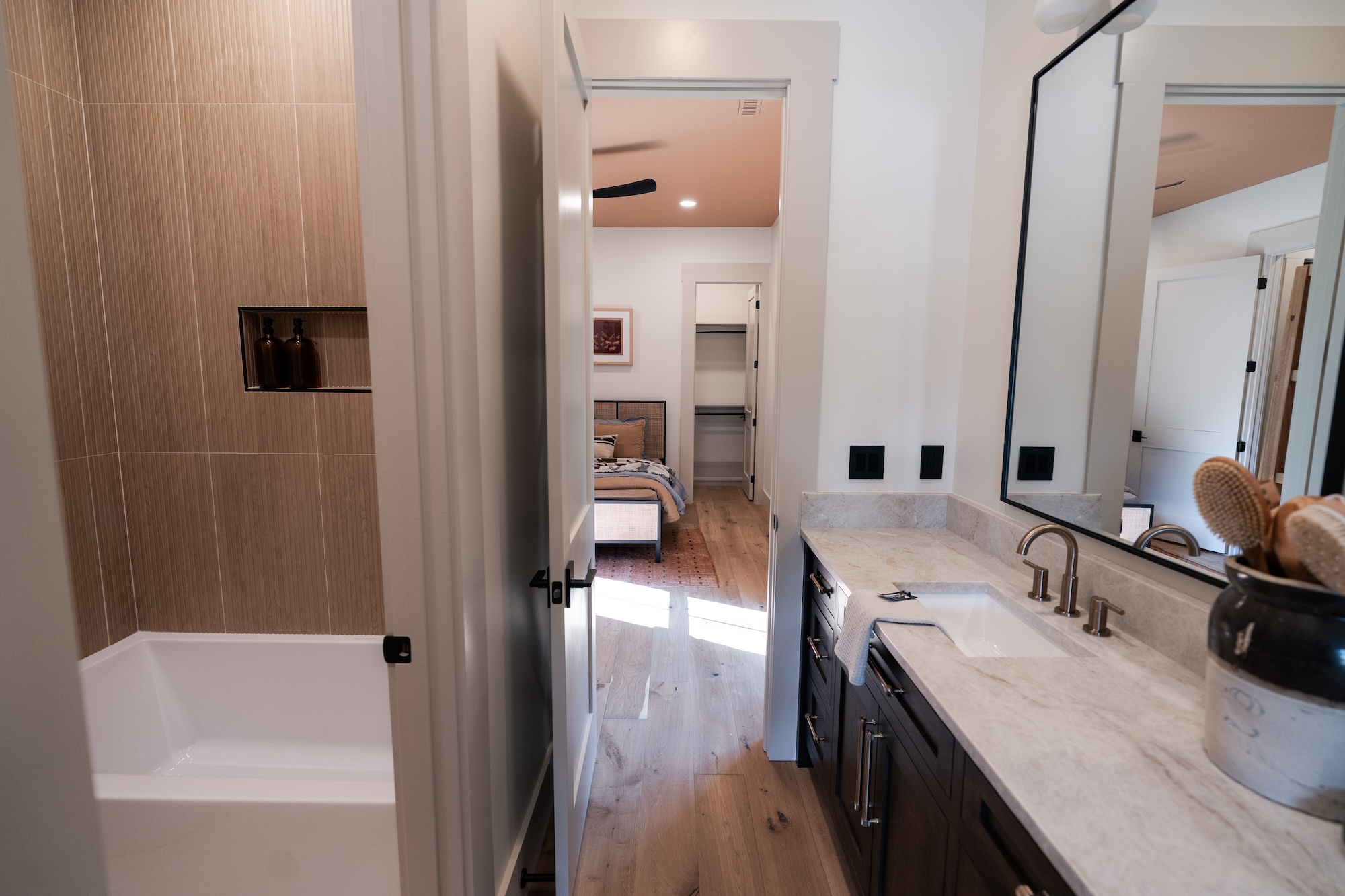 Modern bathroom with a white bathtub on the left featuring a niche with two brown bottles, a marble countertop with a sink and bronze faucet on the right, a large mirror above the sink, and an open door leading to a bedroom with a bed and closet visible.