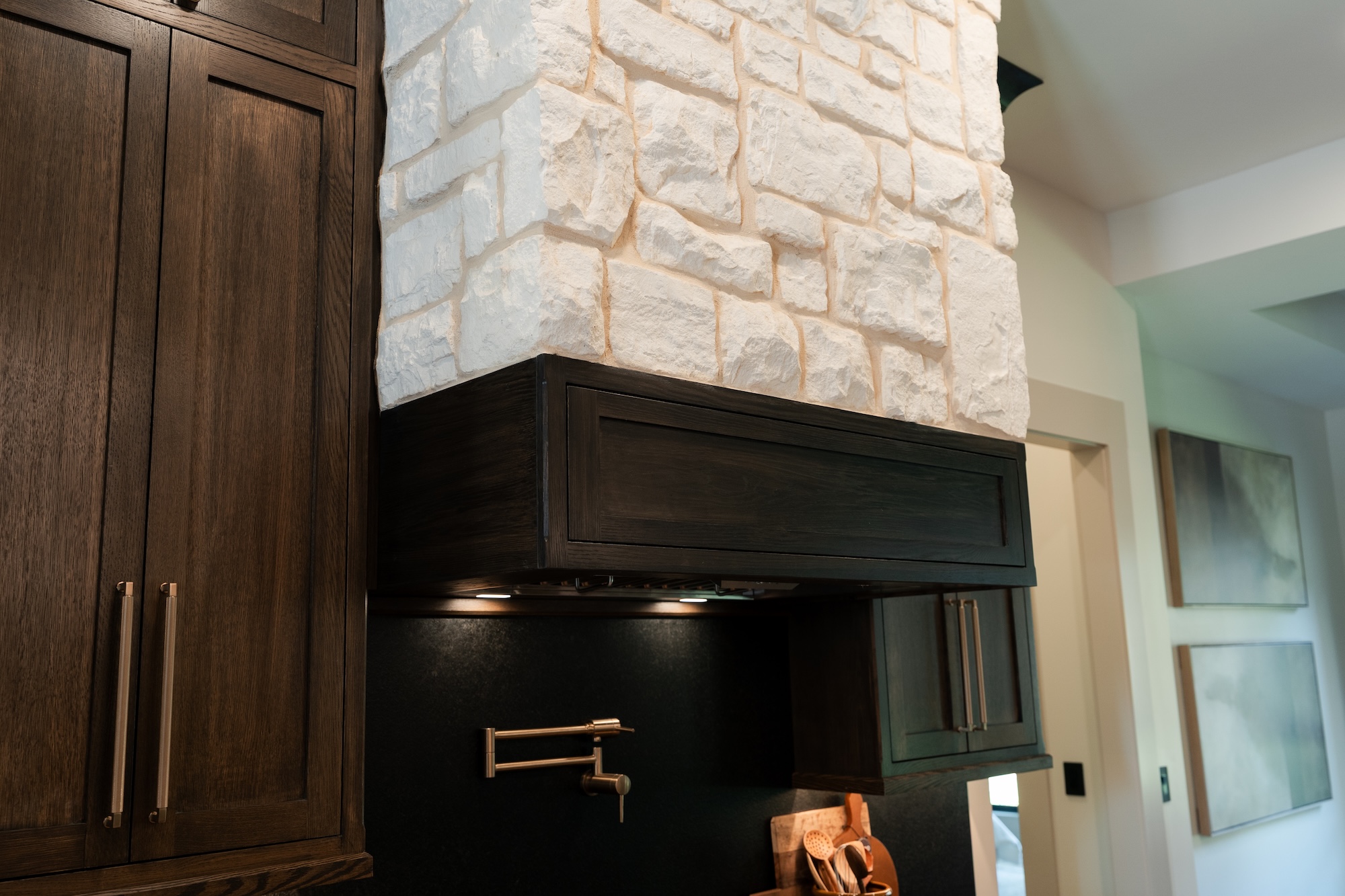 Kitchen corner with dark wooden cabinets and a range hood covered with light textured stone, a brass wall-mounted faucet, and cooking utensils on the counter.