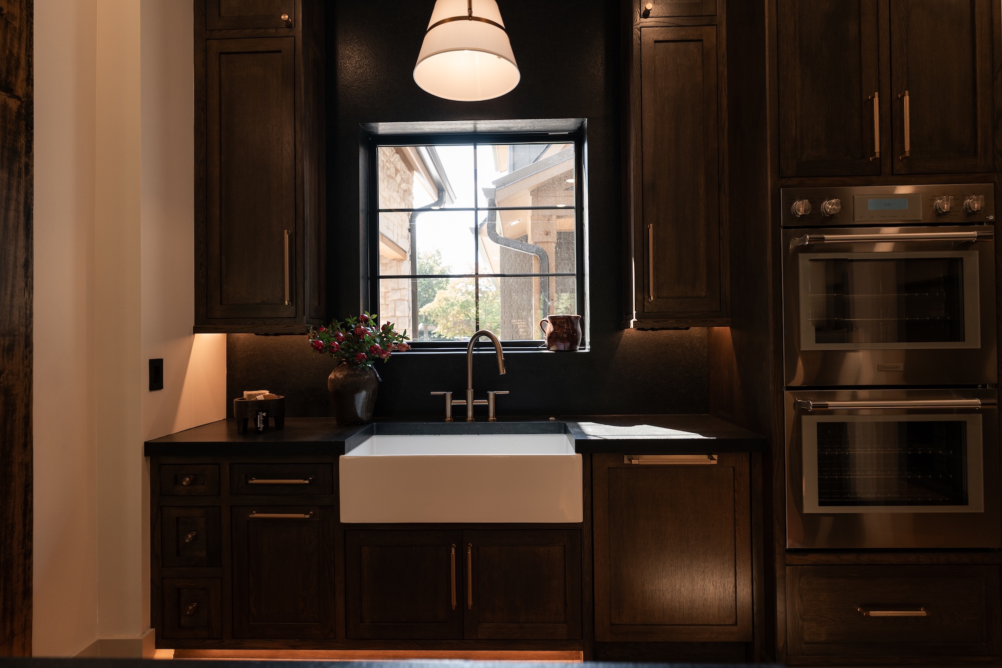 Kitchen with dark wood cabinets, black countertops, and a white apron-front sink beneath a window with black framing, a bronze faucet, a brown vase with red flowers to the left, a small ceramic pitcher on the window sill and double stainless steel ovens to the right under warm pendant light.