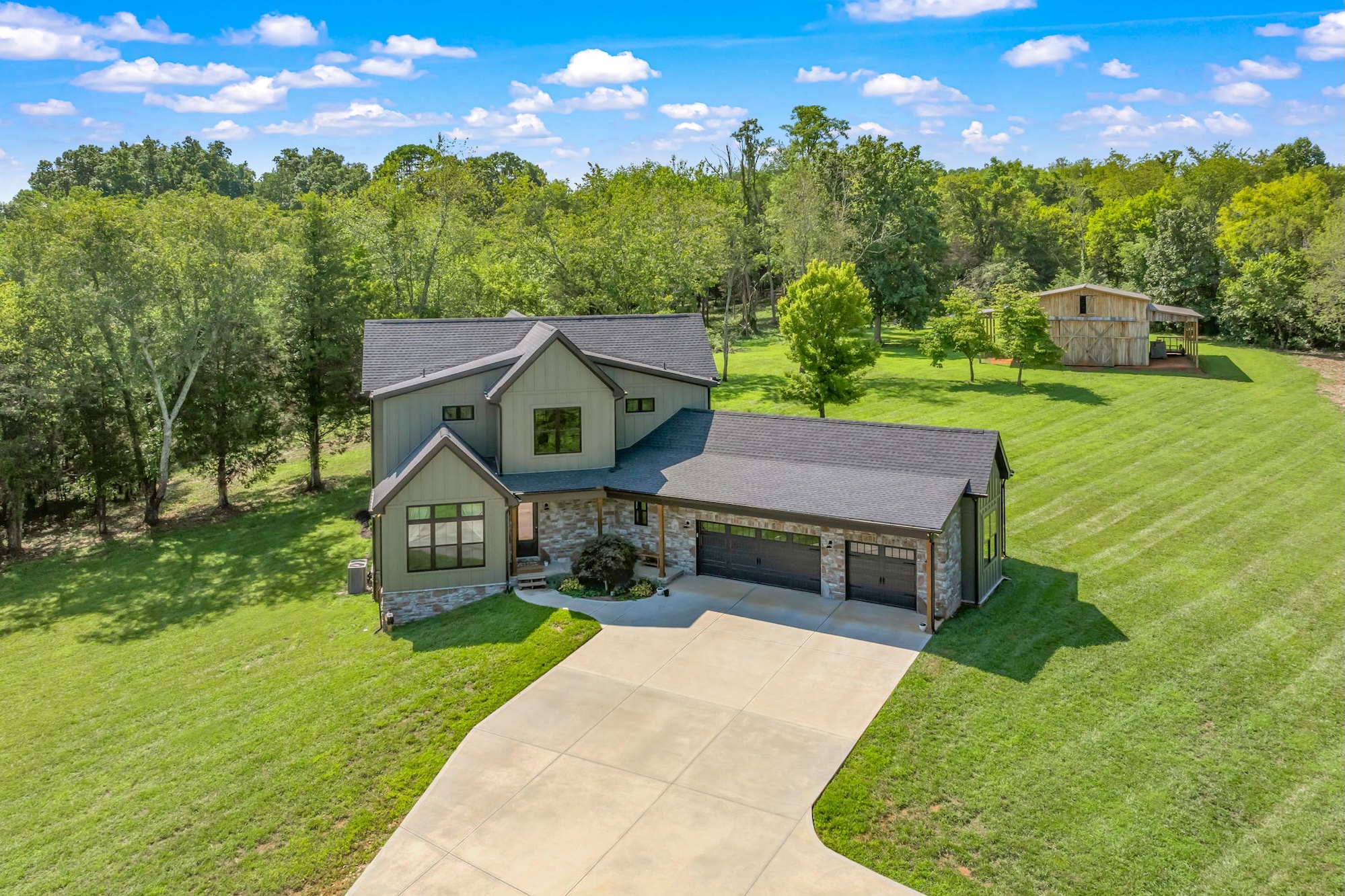 Modern two-story house with stone and siding exterior, a three-car garage, and a wide concrete driveway, surrounded by a large grassy yard with trees and a wooden barn in the background under a partly cloudy blue sky.