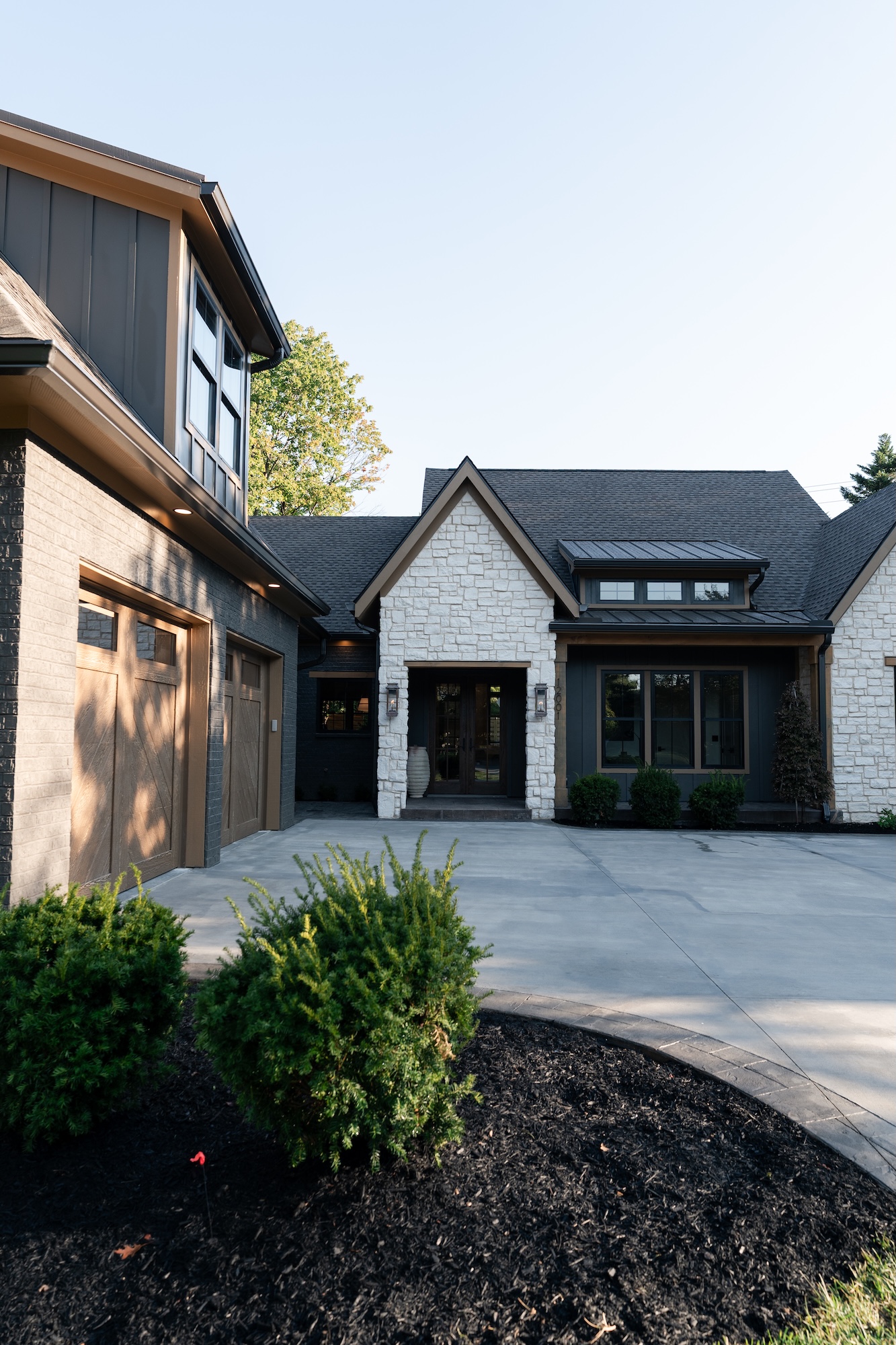 Modern house exterior with stone and dark siding, featuring a covered entryway with lantern-style lights, a garage with wooden doors, and landscaped shrubs in front.